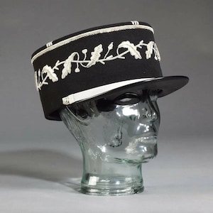 May include: A black hat with a flat brim and a cylindrical crown. The hat has white embroidered floral designs and trim. The hat is displayed on a clear glass head form, highlighting its shape and details. A stylish accessory.