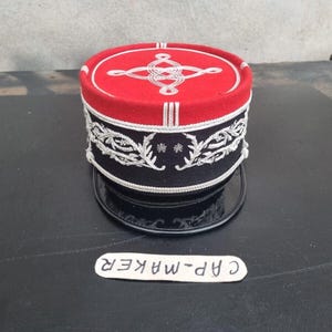 Może przedstawiać: Czapka w stylu wojskowym w kolorze czerwono-czarnym, ozdobiona srebrnym haftem i błyszczącym czarnym rondem. Czapka posiada dekoracyjny wzór na górze i szczegółowe wzory wokół podstawy. Tekst "CAP-MAKER" znajduje się na małej białej etykiecie.