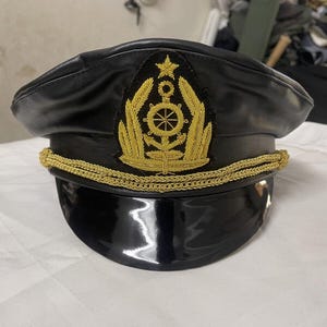 Könnte beinhalten: Schwarze Kapitänsmütze mit glänzendem schwarzem Schirm und goldenem gesticktem Emblem. Das Emblem zeigt einen Stern, ein Steuerrad und Lorbeerblätter. Eine goldene Kordel umrandet die Mütze.
