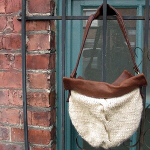 May include: A brown leather and cream knitted tote bag with a long brown leather strap. The bag is hanging on a metal gate.