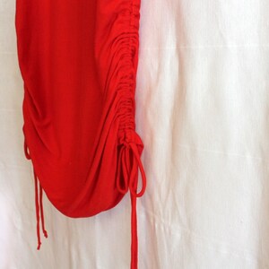 May include: Close-up of a vibrant red garment with a gathered, ruched design along the side. The fabric is smooth and drapes softly, with drawstring details adding texture and visual interest. The background is a plain white.