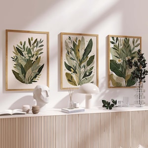 Puede incluir: Tres impresiones artísticas botánicas enmarcadas con follaje verde y beige. Las impresiones se exhiben en una pared blanca sobre una consola blanca. Los marcos son de color marrón claro. Hay objetos decorativos sobre la mesa.