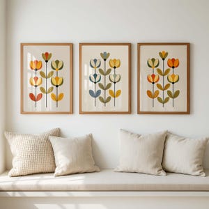 May include: Three framed wall art prints featuring vertical floral arrangements. Each print displays stylized flowers in shades of orange, yellow, olive green, and blue, set against a cream background within light wood frames.
