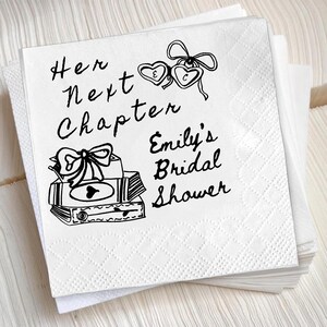 May include: A square white paper napkin with a black ink illustration of a stack of books topped with a bow, two heart-shaped lockets, and the text "Her Next Chapter" and "Emily's Bridal Shower" printed in a handwritten script font.