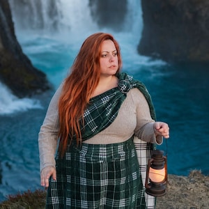 May include: A woman with long red hair stands on a rocky cliff overlooking a waterfall. She is wearing a green and white plaid skirt and a brown sweater. She is holding a lantern in her right hand.
