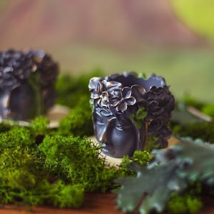 Op de afbeelding: Twee donkergrijze keramische kaarsenhouders met bloemmotieven en een ontwerp van een vrouwengezicht. De kaarsenhouders staan op een bed van groen mos en een houten oppervlak.