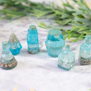 May include: A set of six blue and white resin dice with a beach theme. The dice are shaped like bottles, a pyramid, and a barrel. The dice have white numbers and are filled with glitter and small white shells.