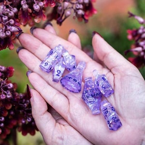 May include: A set of eight purple and clear iridescent dice with black numbers. The dice are in the palm of a hand.