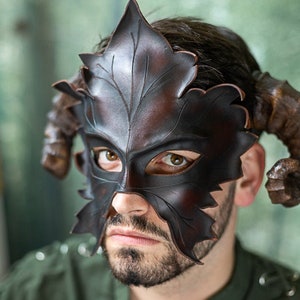Puede incluir: Una m&aacute;scara de cuero marr&oacute;n con un dise&ntilde;o de hoja y cuernos. La m&aacute;scara cubre los ojos y parte de la nariz y las mejillas.