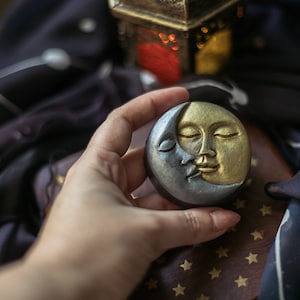 May include: A round soap with a celestial design. The soap is split into two halves: one silver, the other gold, each depicting a serene face of the moon. A vintage-style lantern sits in the background.