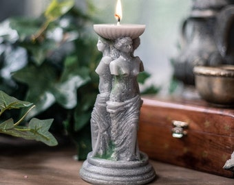 May include: A gray candle shaped like three classical female figures supporting a shallow dish, with a lit flame. The candle is on a round base and has a weathered appearance. The background includes greenery and a wooden box.