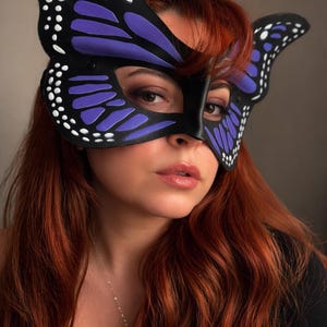 May include: A black butterfly mask with purple and white detailing. The mask features a butterfly wing design and is worn over the eyes. The mask is made of a smooth material, a costume accessory.