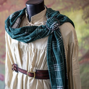 May include: A white linen shirt with a green and white plaid scarf tied around the neck. The scarf is secured with a metal pin. A brown leather belt with a metal buckle is worn around the waist.