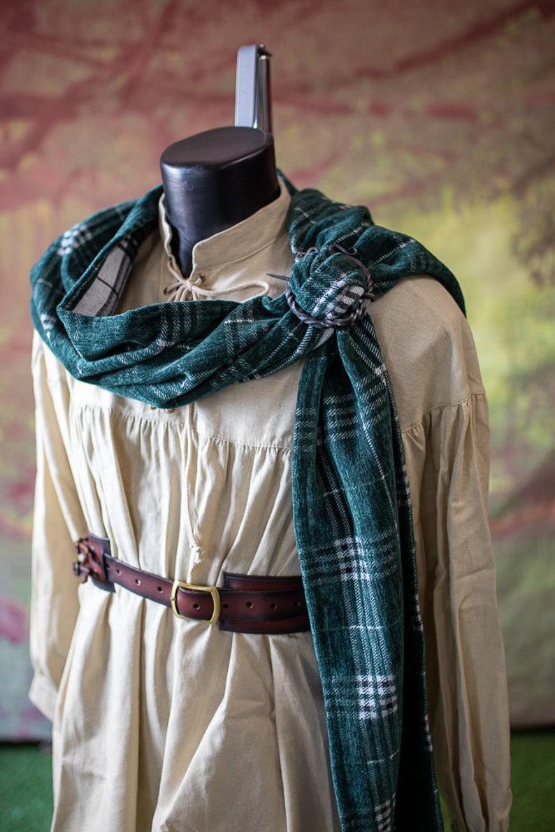 May include: A white linen shirt with a green and white plaid scarf tied around the neck. The scarf is secured with a metal pin. A brown leather belt with a metal buckle is worn around the waist.