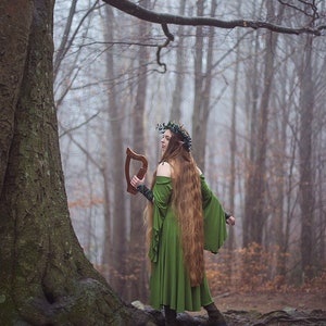 May include: A woman wearing a green dress and a leaf crown holds a wooden harp in a misty woodland setting.