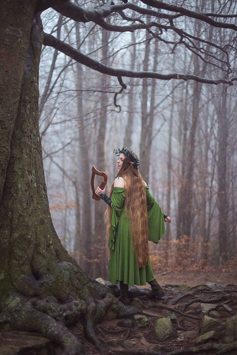 May include: A woman wearing a green dress and a leaf crown holds a wooden harp in a misty woodland setting.