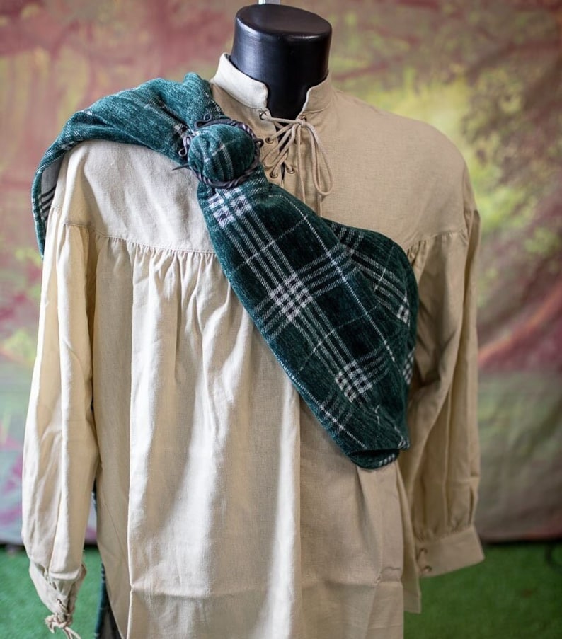 May include: A white linen shirt with a green and white plaid sash worn over the shoulder. The sash is secured with a metal ring.