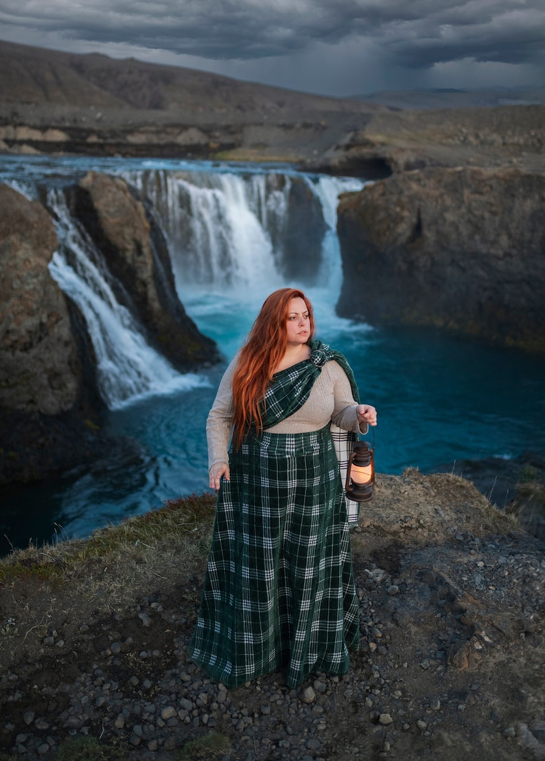 May include: A woman with long red hair stands on a rocky cliff overlooking a waterfall. She is wearing a green and white plaid skirt and a brown sweater. She is holding a lantern in her right hand.
