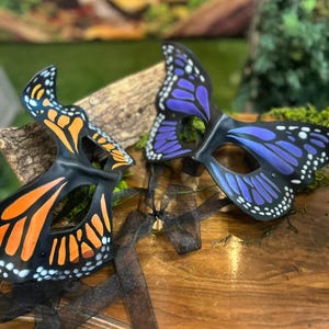 May include: Two butterfly masks with black frames. One mask has orange and black wing patterns, the other has purple and black wing patterns. Both masks have white dots along the edges. Black ribbons are attached to the masks.