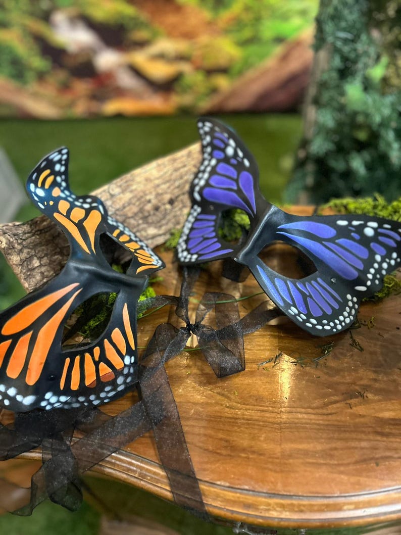 May include: Two butterfly masks with black frames. One mask has orange and black wing patterns, the other has purple and black wing patterns. Both masks have white dots along the edges. Black ribbons are attached to the masks.