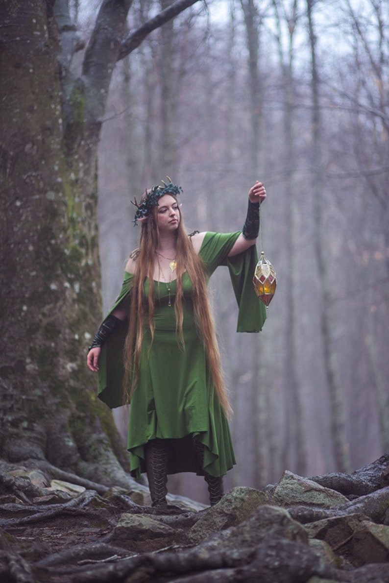 May include: A woman wearing a green dress and a leafy crown holds a stained glass lantern in a woodland setting.