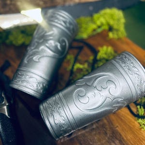 May include: Two silver-colored metal arm cuffs with ornate, embossed floral designs. The cuffs are cylindrical and appear to be made of a durable material. The background includes greenery and a wooden surface, suggesting a rustic or fantasy theme.