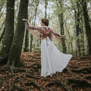 May include: A woman wearing a white dress and a brown and beige feathered wings costume stands in a forest. The forest floor is covered in fallen leaves.
