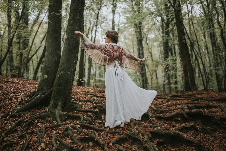 May include: A woman wearing a white dress and a brown and beige feathered wings costume stands in a forest. The forest floor is covered in fallen leaves.