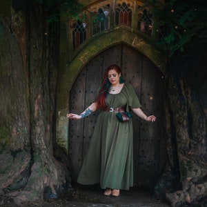 K&ouml;nnte beinhalten: Eine Frau in einem gr&uuml;nen Kleid steht vor einer Holzt&uuml;r mit einem Steinbogen. Sie hat langes rotes Haar mit blauen Str&auml;hnen und tr&auml;gt einen braunen Lederg&uuml;rtel mit einer Tasche.