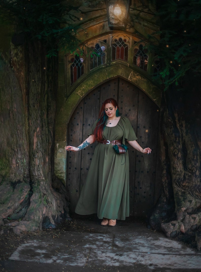K&ouml;nnte beinhalten: Eine Frau in einem gr&uuml;nen Kleid steht vor einer Holzt&uuml;r mit einem Steinbogen. Sie hat langes rotes Haar mit blauen Str&auml;hnen und tr&auml;gt einen braunen Lederg&uuml;rtel mit einer Tasche.