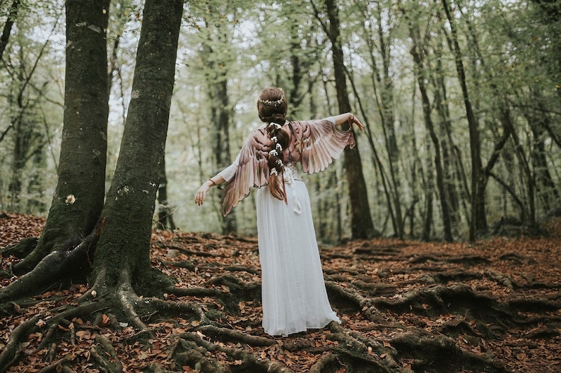 May include: A woman wearing a white dress and a brown feathered shawl stands in a forest. The shawl is shaped like wings and the woman is looking away from the camera.