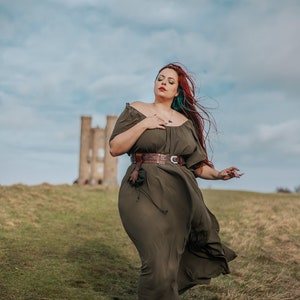 K&ouml;nnte beinhalten: Eine Frau mit roten Haaren und einem gr&uuml;nen Kleid steht auf einem Feld mit einem Steinturm im Hintergrund. Sie tr&auml;gt einen braunen Lederg&uuml;rtel und braune Stiefel.