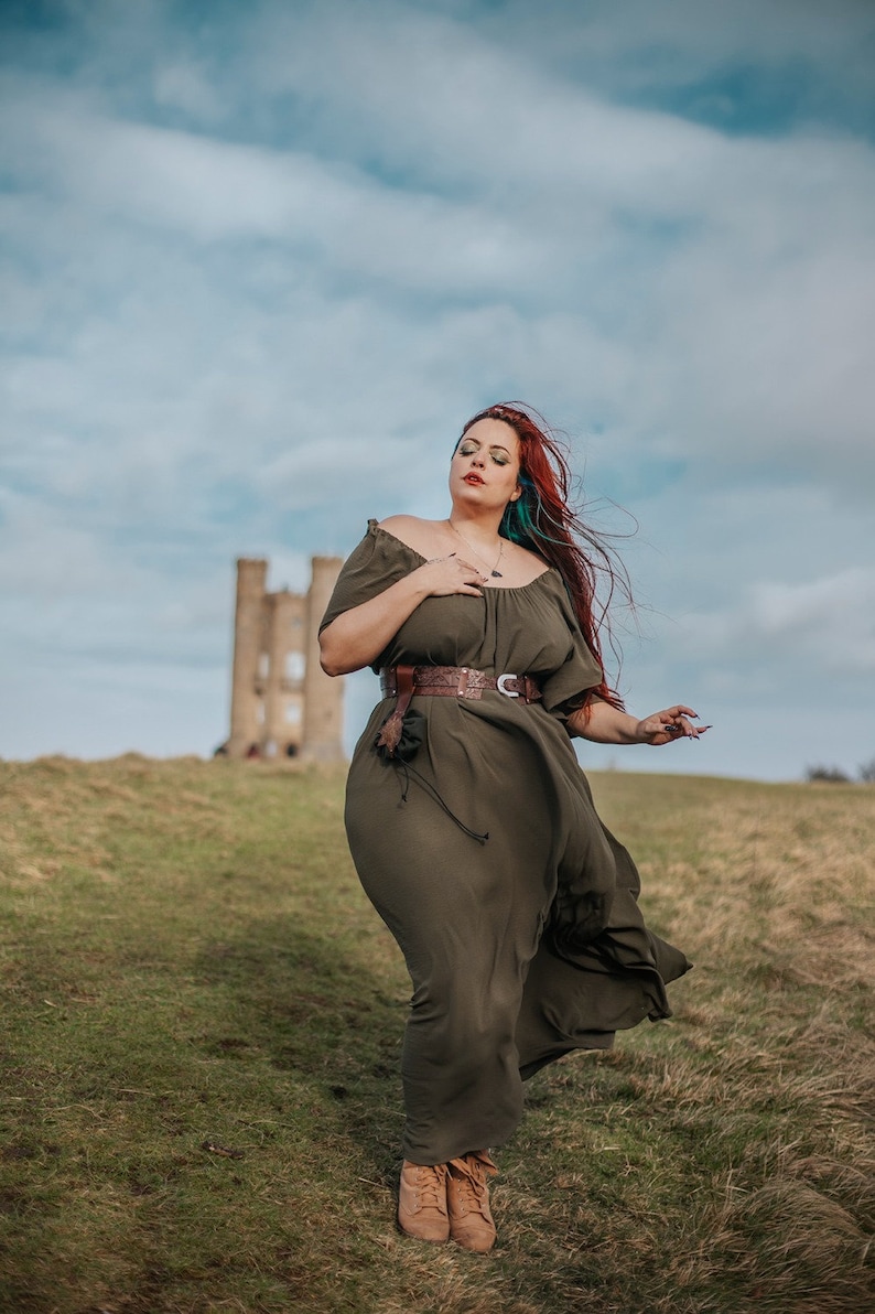 K&ouml;nnte beinhalten: Eine Frau mit roten Haaren und einem gr&uuml;nen Kleid steht auf einem Feld mit einem Steinturm im Hintergrund. Sie tr&auml;gt einen braunen Lederg&uuml;rtel und braune Stiefel.
