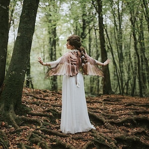 May include: A woman wearing a white dress and a brown and beige feathered wings costume stands in a forest with brown leaves and tree roots on the ground.