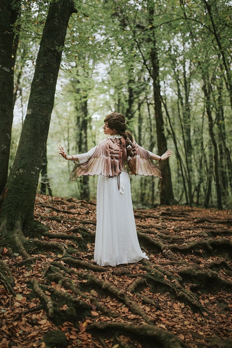 May include: A woman wearing a white dress and a brown and beige feathered wings costume stands in a forest with brown leaves and tree roots on the ground.