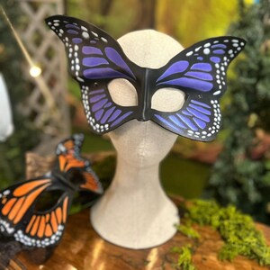 May include: A butterfly mask in black, purple, and white is displayed on a mannequin head. An additional mask with orange and black wings is visible. These masks are likely for costume or decorative use.
