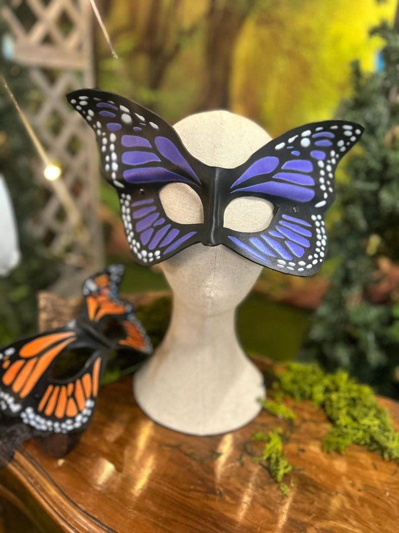 May include: A butterfly mask in black, purple, and white is displayed on a mannequin head. An additional mask with orange and black wings is visible. These masks are likely for costume or decorative use.