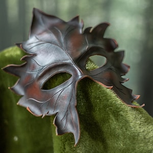 Puede incluir: Una m&aacute;scara de cuero marr&oacute;n con forma de hoja con agujeros para los ojos. La m&aacute;scara est&aacute; sobre una superficie verde y peluda.