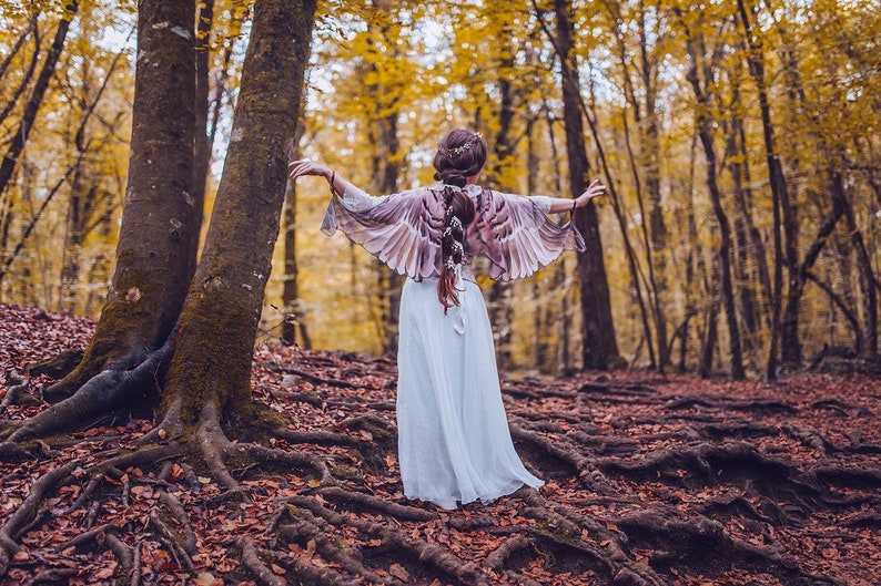 May include: A woman wearing a white dress and a feathered wing shawl stands in a forest with fall foliage. The trees have brown bark and the ground is covered in fallen leaves.
