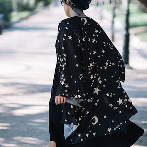 Pode incluir: Um quimono preto com um padrão celestial de estrelas brancas e luas crescentes. O quimono é usado sobre um vestido preto e combinado com botas pretas. A imagem é tirada ao ar livre em um dia ensolarado.
