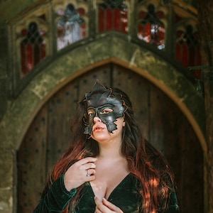 Puede incluir: Una mujer con un vestido de terciopelo verde y una m&aacute;scara de cuero negro con forma de hoja se encuentra frente a una puerta arqueada con vidrieras.