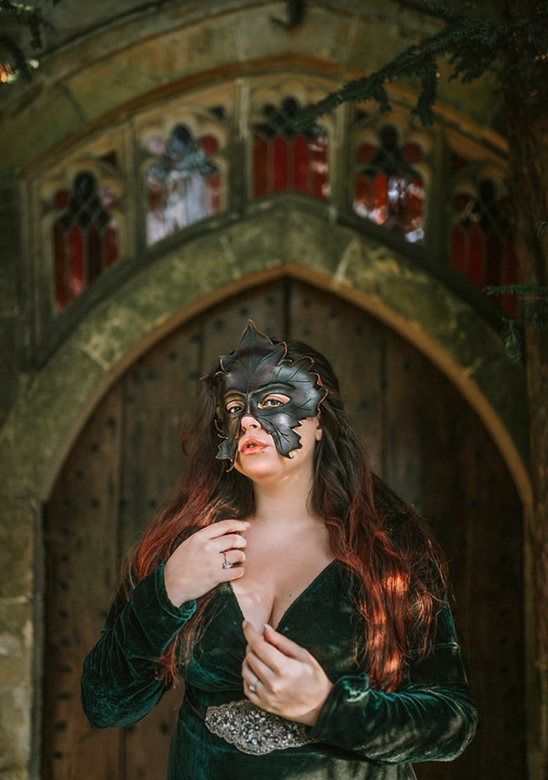 Puede incluir: Una mujer con un vestido de terciopelo verde y una m&aacute;scara de cuero negro con forma de hoja se encuentra frente a una puerta arqueada con vidrieras.
