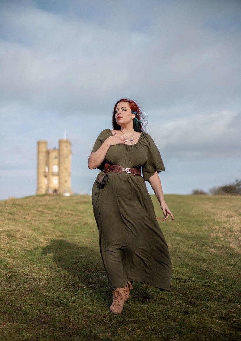 K&ouml;nnte beinhalten: Eine Frau mit roten Haaren und einem gr&uuml;nen Kleid steht auf einem grasbewachsenen H&uuml;gel mit einem Steinturm im Hintergrund.