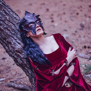 Puede incluir: Una mujer con un vestido de terciopelo rojo oscuro y una m&aacute;scara con forma de hoja sobre los ojos. Se apoya en el tronco de un &aacute;rbol en un entorno de bosque.