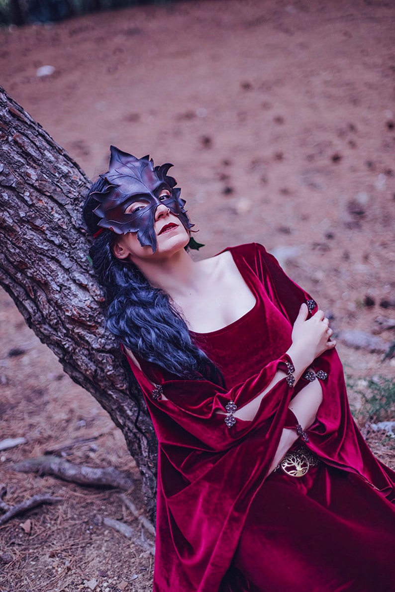 Puede incluir: Una mujer con un vestido de terciopelo rojo oscuro y una m&aacute;scara con forma de hoja sobre los ojos. Se apoya en el tronco de un &aacute;rbol en un entorno de bosque.