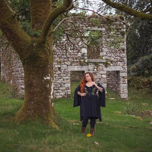 May include: A woman with long red hair stands in front of a stone building ruin. She is wearing a black dress with a green and black patterned bodice and black boots.