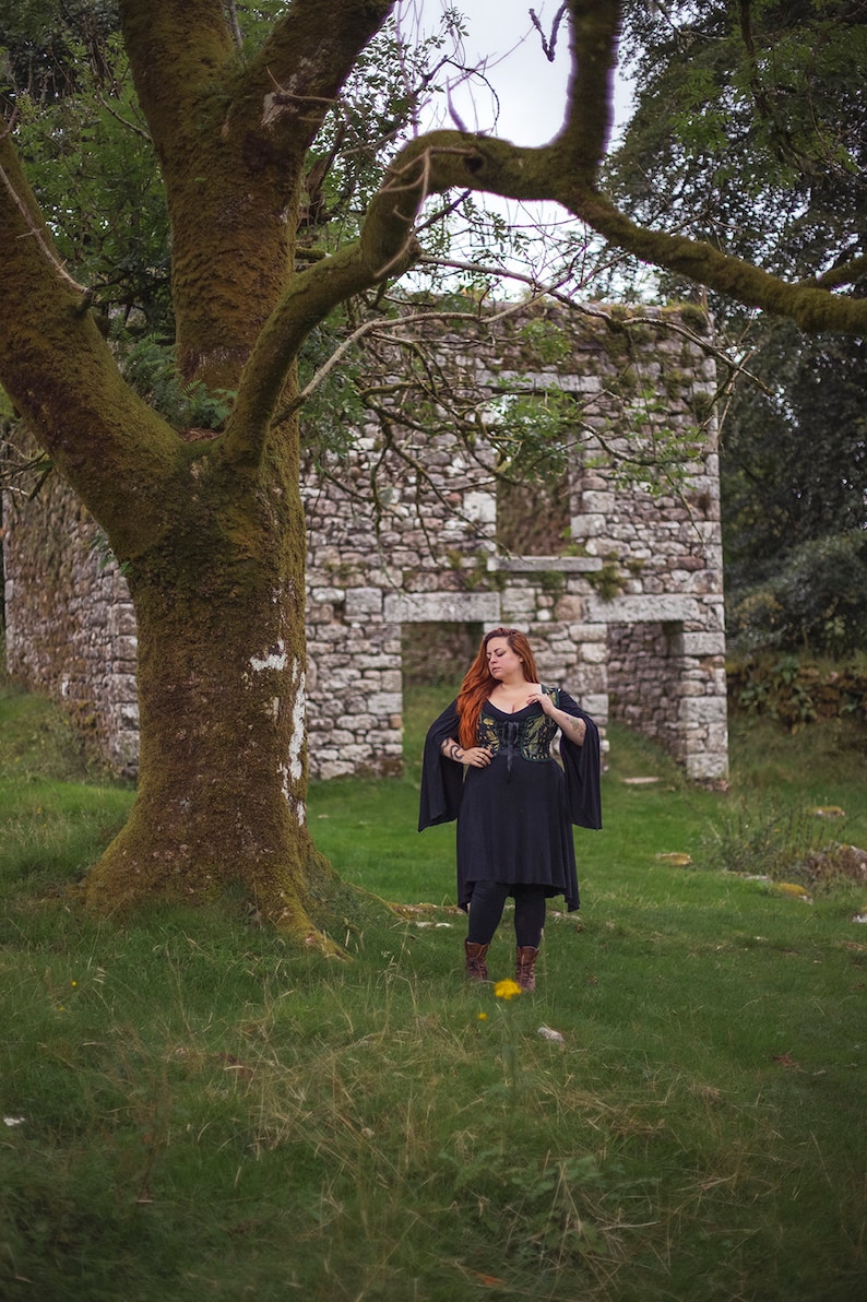 May include: A woman with long red hair stands in front of a stone building ruin. She is wearing a black dress with a green and black patterned bodice and black boots.