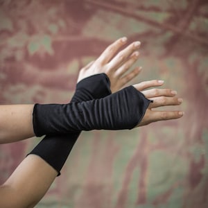 Puede incluir: Se muestran guantes negros sin dedos, un accesorio elegante. Los guantes están hechos de una tela oscura y suave y cubren los antebrazos, dejando los dedos expuestos. El fondo es una mezcla de colores apagados y texturizados.