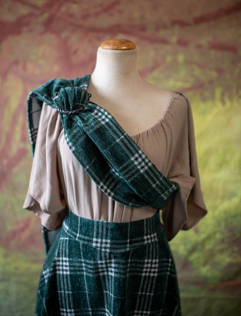May include: A green and white plaid skirt with a matching plaid scarf draped over a beige blouse. The scarf is secured with a metal ring.