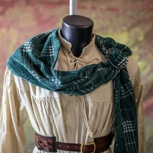 May include: A cream-colored shirt with a green and white plaid scarf. The shirt has a lace-up neckline and a brown leather belt.